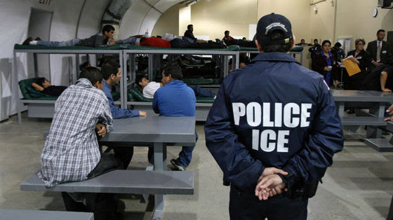 an I.C.E. office standing guard over migrants sitting at a table in a detention center